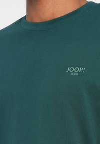 Tamsiai žalia trumpomis rankovėmis marškinėliai, pagaminti iš minkštos medvilnės, su mažesniu baltonu "JOOP! JEANS" logotipu krūtinėje. Lygus paviršius, apvalaus kaklo dizainas.