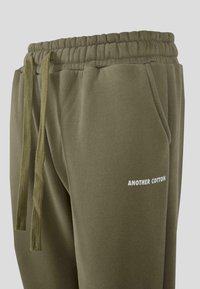 Olivenfarvede sweatpants med elastisk talje, snorbinding, sidelommer og lille hvid "ANOTHER COTTON" tekst på venstre lår.