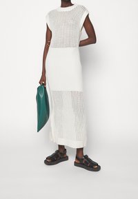 Gebreide witte jurk met korte mouwen en open geweven delen. Gecombineerd met zwarte sandalen en een groene, gevouwen clutch.