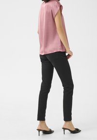 Blouse rose sans manches avec une texture lisse, associée à un jean noir ajusté. Le mannequin porte des mules à talons noires avec un accent décoratif.
