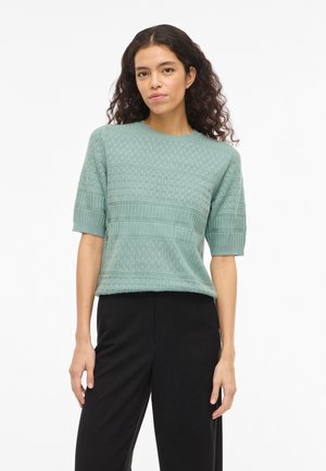 Žena s dlhými kučeravými vlasmi nosí svetlozelené textúrované pletené tričko s krátkym rukávom a čierne nohavice na bielom pozadí.