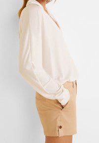 Blusa de tono crema de manga larga con escote en V y ajuste holgado, combinada con pantalones cortos color canela que cuentan con bolsillos laterales y detalles de botones.