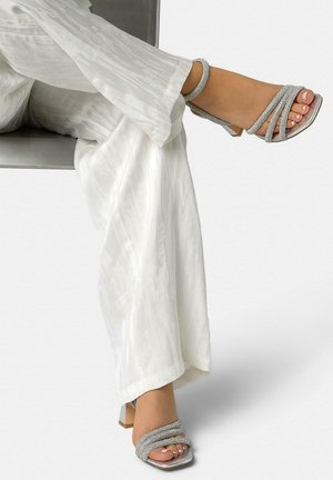 Gekreuzte Füße, die silberne Riemchen-Sandaletten mit Blockabsatz tragen, kombiniert mit weißen, strukturierten, weit geschnittenen Hosen vor einem schlichten Hintergrund.