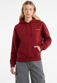 Burgundská mikina s kapucňou a predným vreckom, nastaviteľnými šnúrkami a bielym logom "Guess Jeans". Mäkkučký materiál s rebrovanými manžetami.