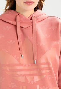 Rosa hoodie med ett mönster av små logotyper, en stor logotyp på framsidan och snören med silverändar. Mjuk material och avslappnad passform.