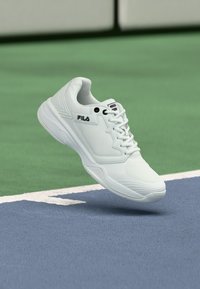 Valge FILA spordijalats, millel on paelad, hõljuv sinise ja rohelise tennisepinna kohal valge piirijoone lähedal.