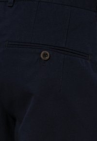 Poche arrière d’un pantalon bleu marine foncé avec une fermeture à un bouton et un détail de poche passepoilée horizontale.