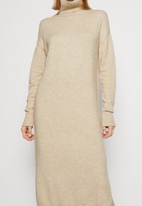 Pletené béžové šaty s vysokým límcem, dlouhými rukávy, žebrovanými manžetami a volným střihem. Měkká textura a střední délka, minimalistické a bez švů.