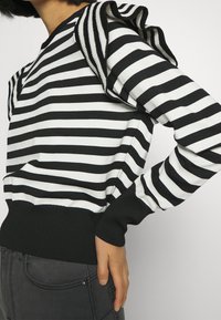 Maglione a righe bianche e nere con scollatura a costine e ampia rouches accentuata sulla spalla. Polsini e orlo aderenti.