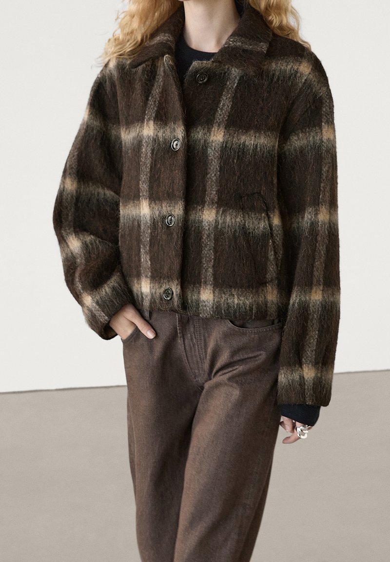 Personne portant une veste boutonnée marron à carreaux duveteuse et un pantalon marron assorti, avec une main dans la poche et des bagues argentées aux doigts.