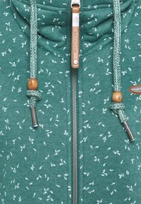 Felpa verde con zip e motivo a foglie, caratterizzata da un tessuto testurizzato, dettagli in legno e lacci intrecciati.