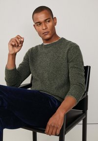 Mann in grünem Strickpullover und dunkelblauen Hosen sitzt auf einem schwarzen Stuhl und schaut nachdenklich zur Seite, während er eine Hand nahe seinem Gesicht hebt.