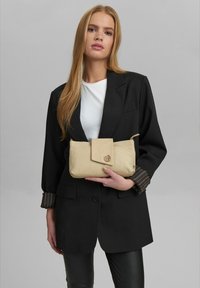 Borsa a tracolla in nylon beige con chiusura a patta, logo in rilievo e superficie liscia, abbinata a un blazer nero e maniche a righe.