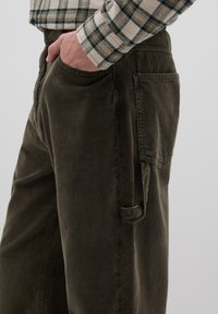 Dunkelolivfarbene Cordhosen mit glatter Textur, ausgestattet mit Seitentaschen und einem kleinen Riemendetail am Bund.