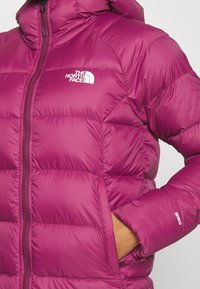Personne portant une veste matelassée magenta The North Face avec une main dans la poche latérale et le logo visible sur la poitrine et la manche.