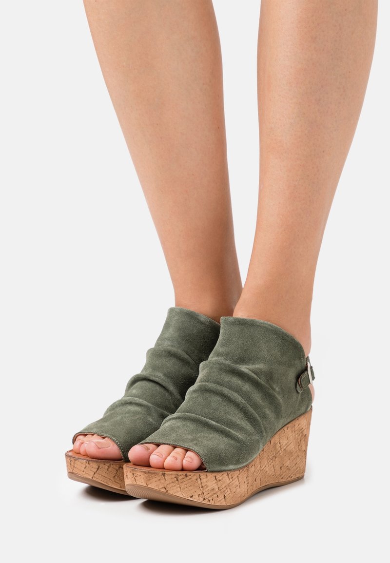 Sandales compensées en daim vert avec orteils ouverts, présentant une plateforme en liège texturé et un design en tissu froncé. Détail de boucle réglable sur le côté.