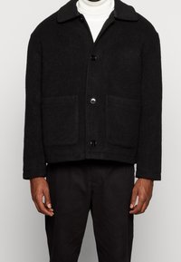 Giacca nera in lana con design a colletto, due tasche frontali e chiusura con bottoni. Tessuto liscio e vestibilità leggermente oversize.