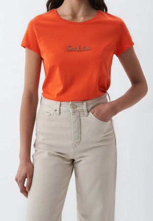 Femme portant un t-shirt orange vif à manches courtes avec le texte "Salsa" et un jean taille haute beige, une main dans la poche sur un fond uni.