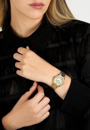 Orologio da polso color oro con quadrante rotondo, quadrante bianco, numeri romani e cinturino metallico con maglie di dimensioni varie. Indossato su una mano.