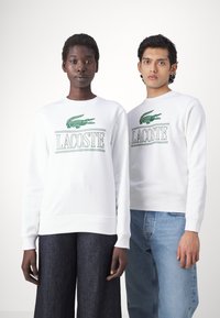 Felpe in cotone bianco con logo verde Lacoste, scollatura rotonda, polsini a costine e vestibilità rilassata, abbinate a denim scuri e chiari.