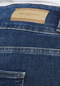 Vaqueros de denim con parche de cuero color beige etiquetado "ONLY CARMAKOMA" cosido encima del bolsillo trasero en tela de color azul oscuro.