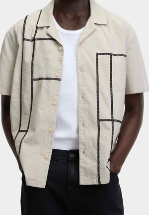 Un homme porte une chemise beige à manches courtes boutonnée avec des lignes noires en grille sur un débardeur blanc côtelé, associée à un pantalon noir.