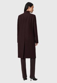 Casaco longo bordeaux com corte reto, gola alta e ventral central nas costas, emparelhado com calças slim combinando. Textura de tecido suave.