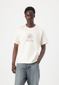 Cream-colored cotton t-shirt with a round neckline. Features a central graphic design in maroon that includes text and decorative elements.