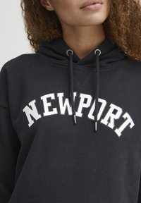 Felpa nera con tasca a canguro, caratterizzata dalla scritta "NEWPORT" in bold bianche. Presenta un cappuccio con cordini e dettagli a costine nel colletto.