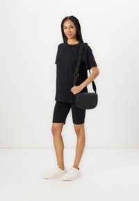 Čierne oversized tričko, čierne bikerské šortky a biele tenisky. Model drží čiernu crossbody tašku s texturovaným logom. Biele pozadie.