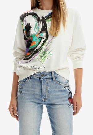 Kvinde iført hvid sweatshirt med farverigt abstrakt ansigtsdesign og lyseblå jeans med små tegneserie- og håndskrevne tekstdetaljer.