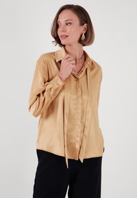 Satin, dugih rukava, svijetlo zlatna bluzica s ovratnikom i vezanim detaljem sprijeda. Ima manžetne na dugmad i gladak materijal. Crne hlače su vidljive.