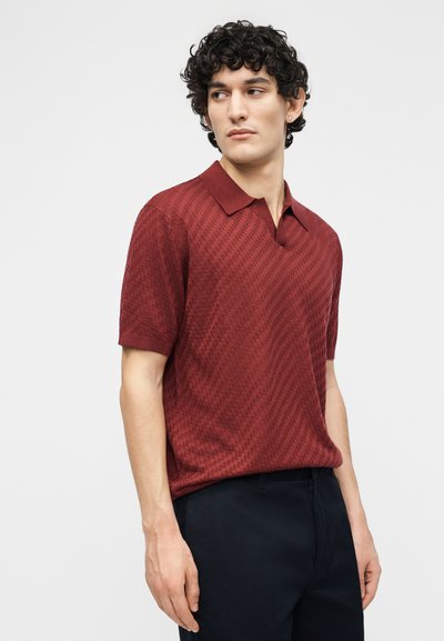 Νέος με κατσαρά μαλλιά που φοράει μπλουζάκι πόλο με κοντά μανίκια σε υφή μπορντό χρώματος και σκούρο παντελόνι, κοιτάζοντας πλάγια σε απλό φόντο.