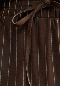 Bruin stof met witte verticale strepen, voorzien van een elastische tailleband, trekkoordsluiting en een zachte textuur. Inclusief een zijzak detail.