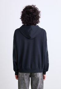 Sort oversized hoodie med snor i hætten, ribbede mansjetter og et grafisk design langs ærmerne, sat sammen med lysegrå distressed jeans.