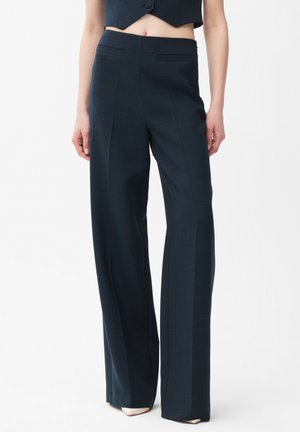 Kobieta ubrana w ciemnogranatowe spodnie o wysokim stanie i szerokich nogawkach, w połączeniu z krótką czarną kamizelką, stojąca na tle białego tła.