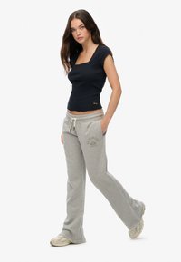 Czarny dopasowany top z falistymi brzegami, w połączeniu z jasnoszarymi dresami z nadrukiem logo. Modelka stoi pewnie, z rękami w kieszeniach.