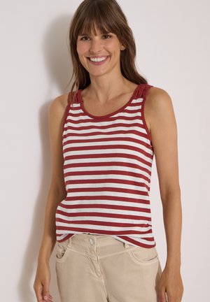 Femme souriante aux cheveux bruns, portant un haut sans manches rayé rouge et blanc et un pantalon beige clair, debout contre un mur uni.