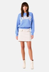 Sudadera azul claro con el texto "L'Amore", combinada con una falda corta blanca con bolsillos delanteros. Llevada con botas blancas hasta el tobillo.