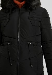 Manteau d'hiver matelassé noir avec fermeture éclair devant, cordons réglables à la taille, poches poitrine à boutons-pression et grand col en fausse fourrure noire.