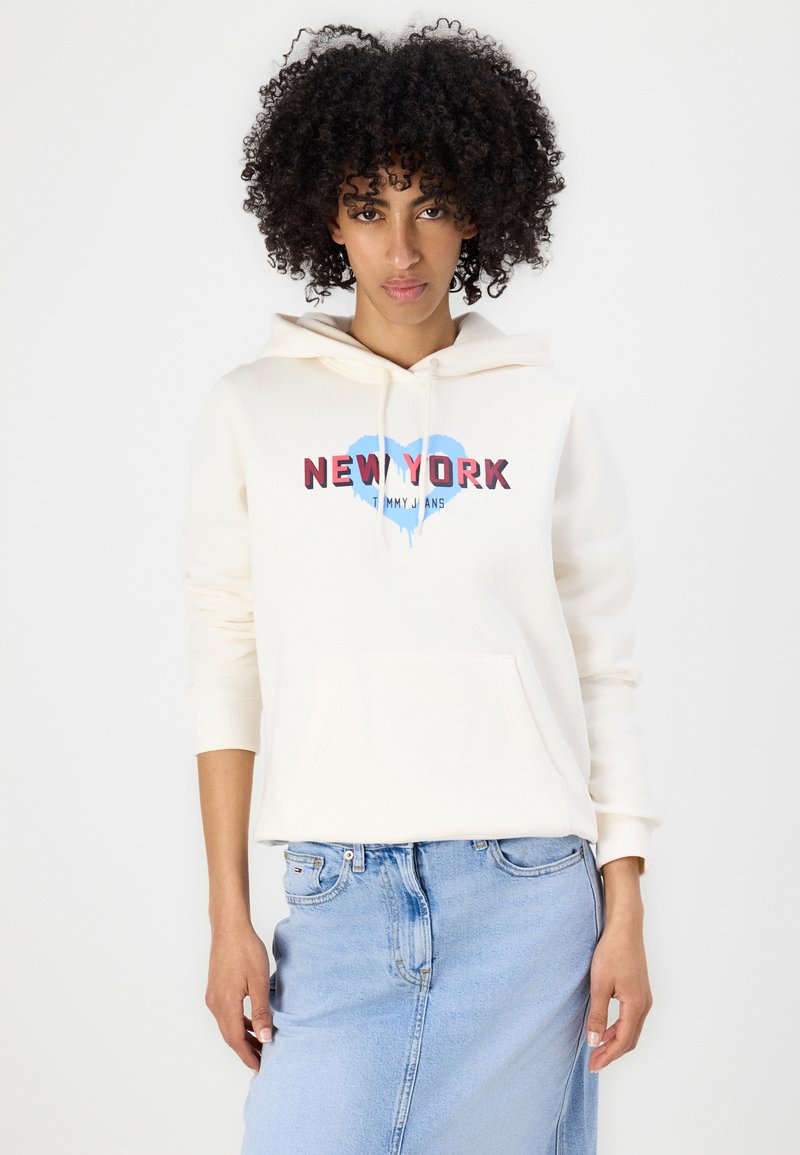 Helles Creme-farbenes Hoodie mit Fronttasche, das „NEW YORK“ in rotem und blauem Grafikdesign zeigt. Kombiniert mit einem hellblauen Jeansrock.