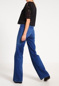 Person som bär blå vida jeans och en svart kortärmad topp med spetsdetaljer, går framför en enkel vit bakgrund.