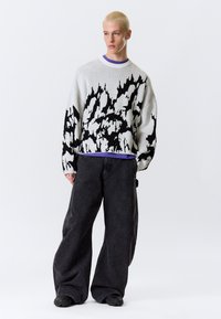 Pull tricoté blanc avec motif abstrait noir, muni d'un col rond et de manches longues ; porté avec un jean noir ample et des chaussures foncées.