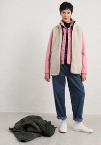 Beige fleece vest over een roze overhemd, gedragen met brede blauwe broek en witte sneakers, met een groene jas op de vloer.