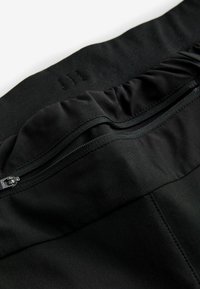 Pantalons de sport noirs avec une texture lisse, une poche zippée et une taille élastique avec trois lignes de couture verticales.