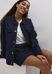 Temno modra jeans jakna s srebrnimi žebljički, dvema žepoma na prsih in kovinskimi gumbi. V kombinaciji z usklajeno denim krilom. Pod njo je bela majica.