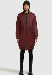 Bordeaux gewatteerde jas met een ritssluiting aan de voorkant, zijzakken, geribde manchetten en een logopatch op de mouw. Draagt over zwarte leggings en laarzen.