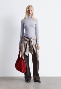 Hellblaues Langarmoberteil, karierte Bluse um die Taille gebunden, dunkelbraune weite Hosen und eine rote Wildleder-Schultertasche. Schwarze Schuhe runden das Outfit ab.