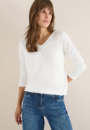Weißes langärmliges Oberteil mit V-Ausschnitt und gerafftem Detail auf der Vorderseite, kombiniert mit blauen Jeans aus Denim. Einfacher, glatter Stoff und lockere Passform.