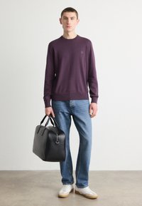 Maglione lavorato a maglia viola con colletto rotondo, jeans blu in denim, scarpe da ginnastica bianche e una borsa da viaggio nera in pelle con doppi manici.